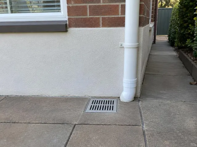 Outdoor gully with grate on dry paving beside a brick house wall and downpipe in Rydalmere