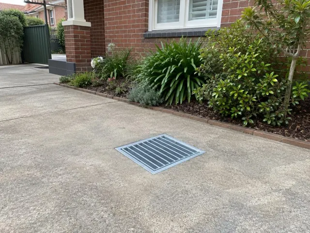 Stormwater driveway grate set in concrete beside brick house and garden beds in Westmead